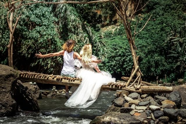 9 Regalos ecológicos para agregar a su lista de regalos de bodas, a girl and a bride sitting on a log bridge above a stream in the woods.